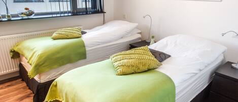 A room with two single beds, each with a green blanket and pillow, a window with blinds, and two abstract paintings on the wall.