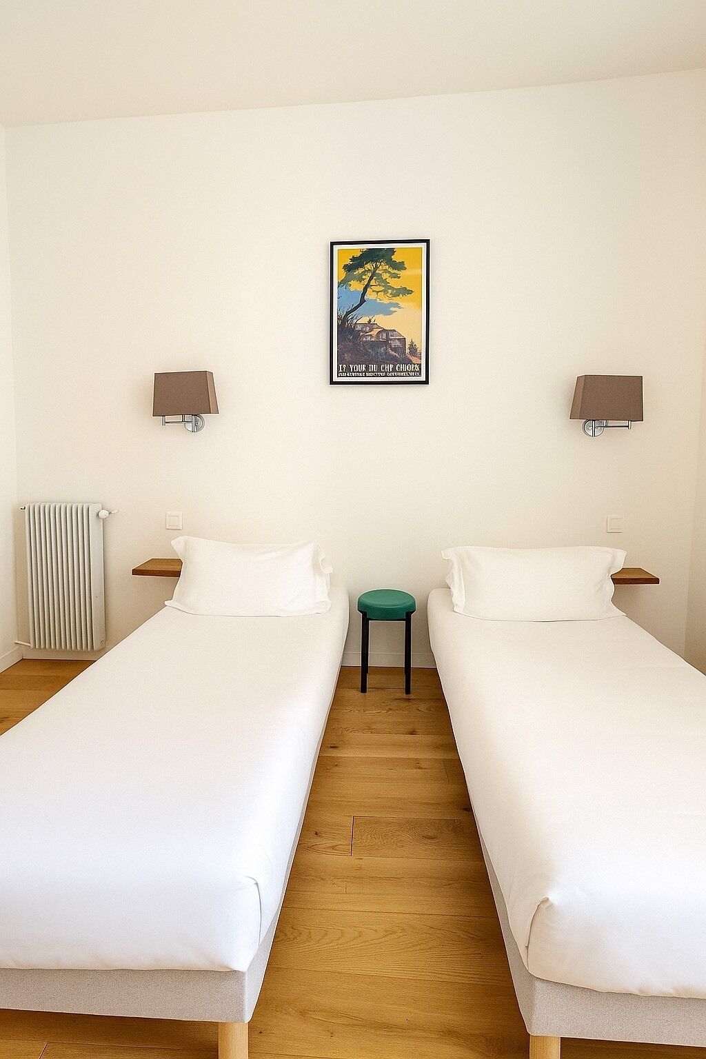 A hotel room with two single beds, a radiator, a small stool, and two wall-mounted lamps.