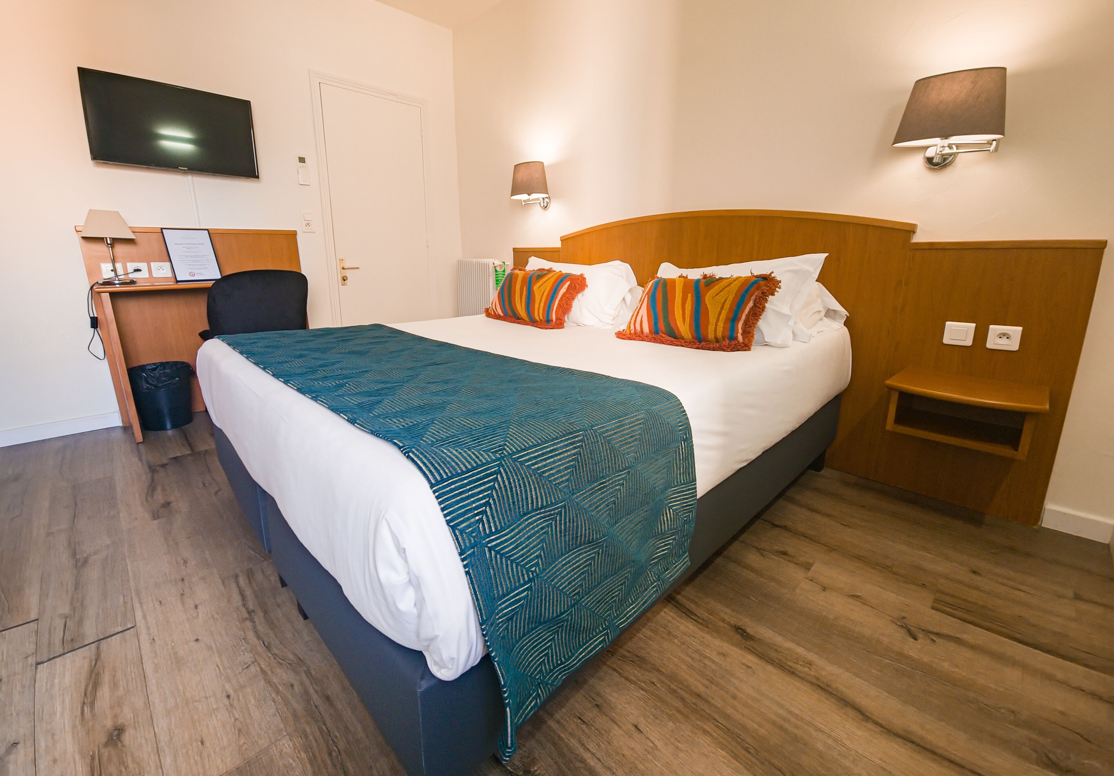 Une chambre d’hôtel équipée d’un lit, d’un bureau, d’une chaise, d’une télévision et d’une lampe fixée au mur.