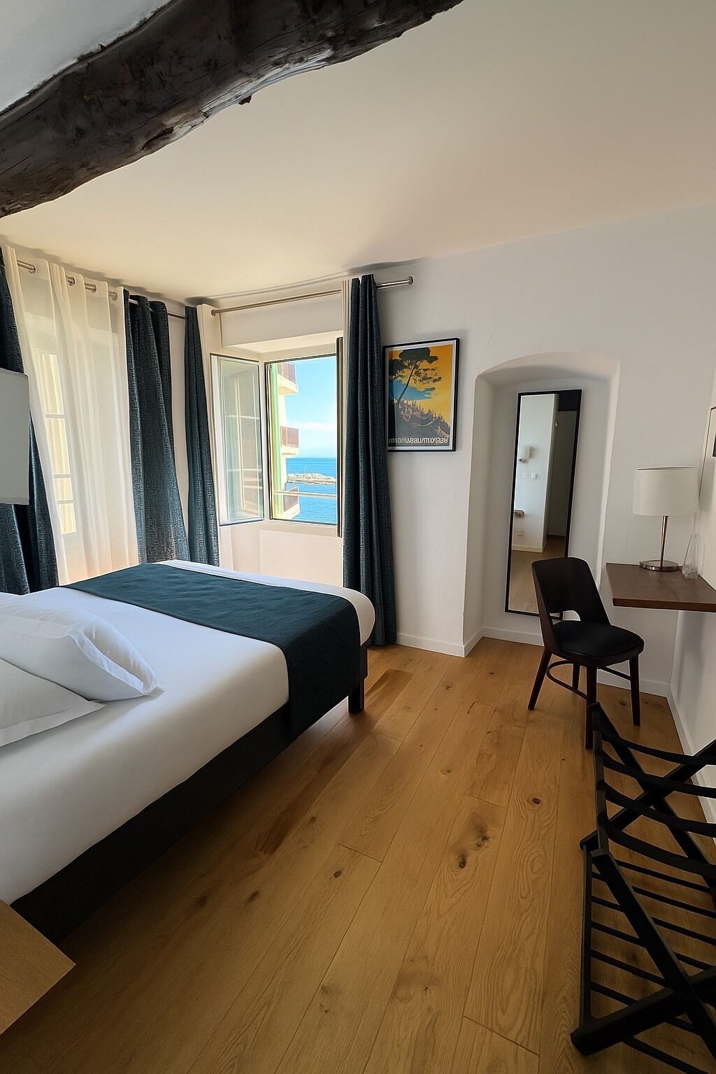 A hotel room with a bed, a desk, a chair, a large window with a view, and a painting on the wall.
