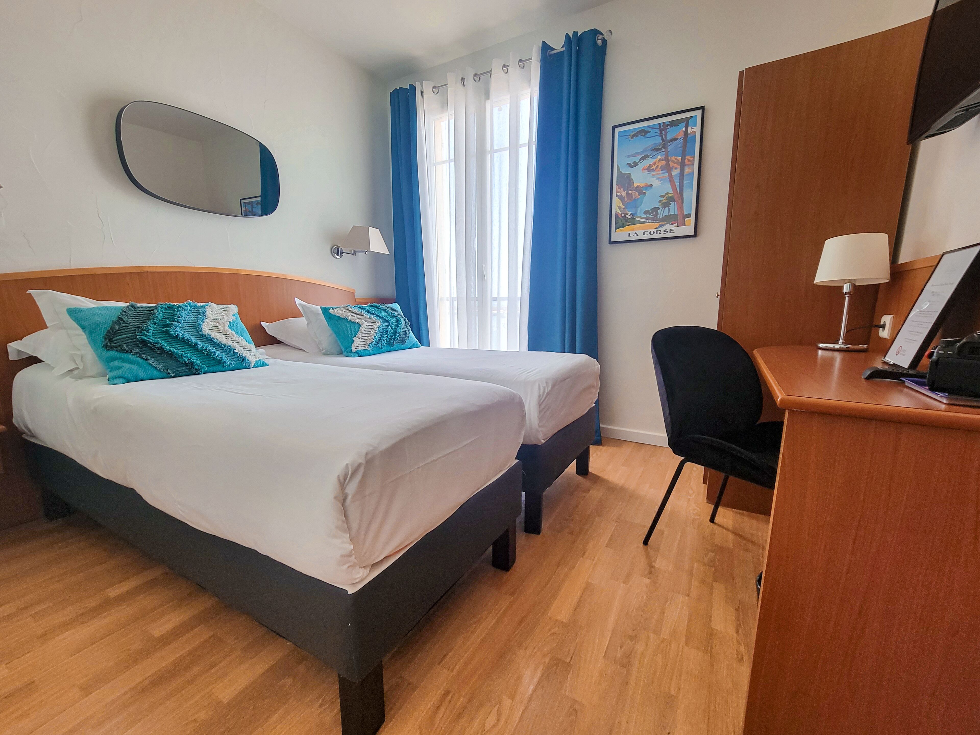 Una habitación de hotel con dos camas, un escritorio, una silla, un espejo, una lámpara y una ventana con cortinas.