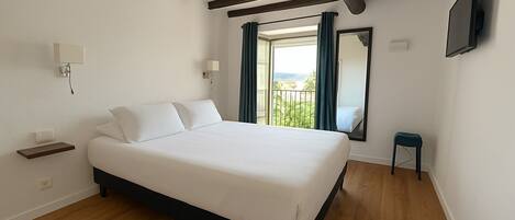 Un dormitorio con cama, vigas de madera, una ventana con cortinas, un taburete pequeño y un televisor de pantalla plana.
