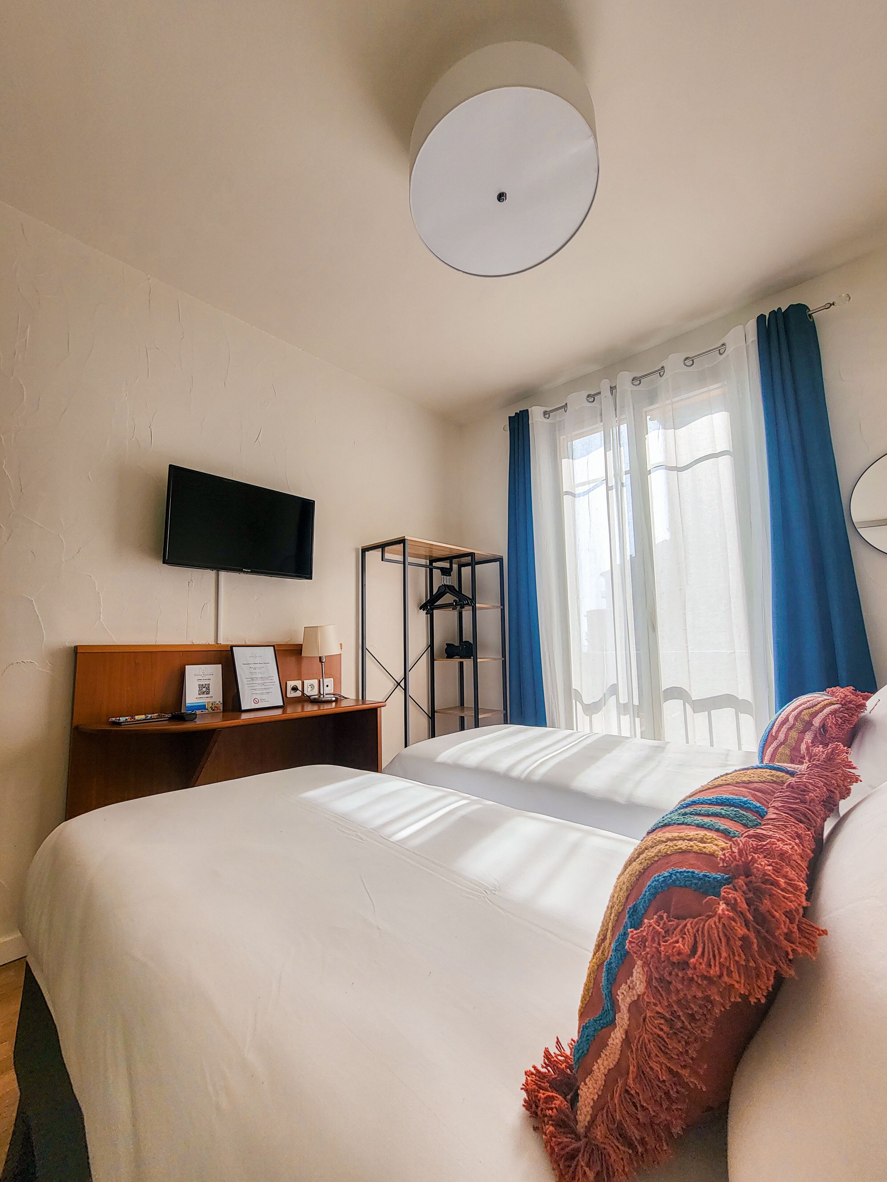 Une chambre d’hôtel avec un lit, un téléviseur fixé au mur, un bureau avec une lampe et une fenêtre avec des rideaux.
