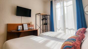 Une chambre d’hôtel avec un lit, un téléviseur fixé au mur, un bureau avec une lampe et une fenêtre avec des rideaux.