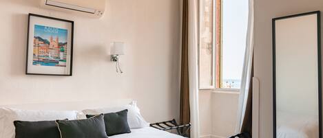 Habitación de hotel con cama, un cuadro enmarcado de un paisaje urbano, aire acondicionado en la pared, una ventana con cortinas y una silla.