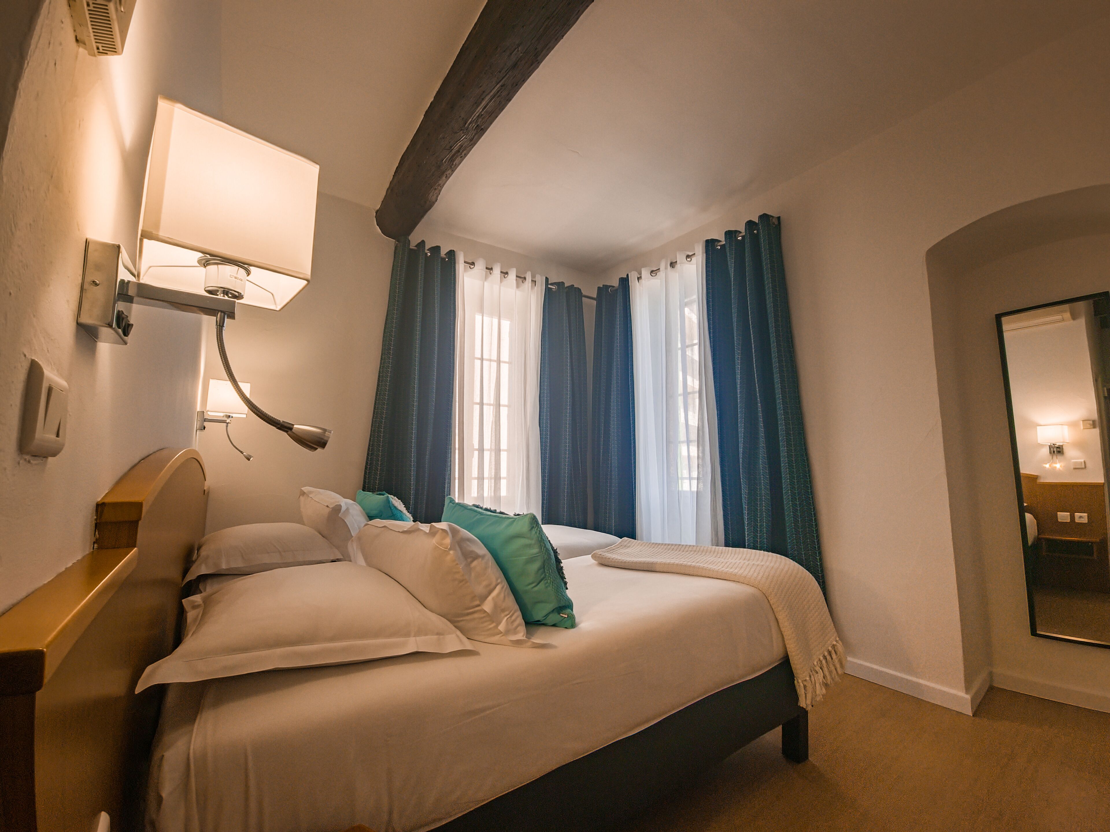 Una habitación de hotel con cama, lámpara de noche, espejo y ventana con cortinas.