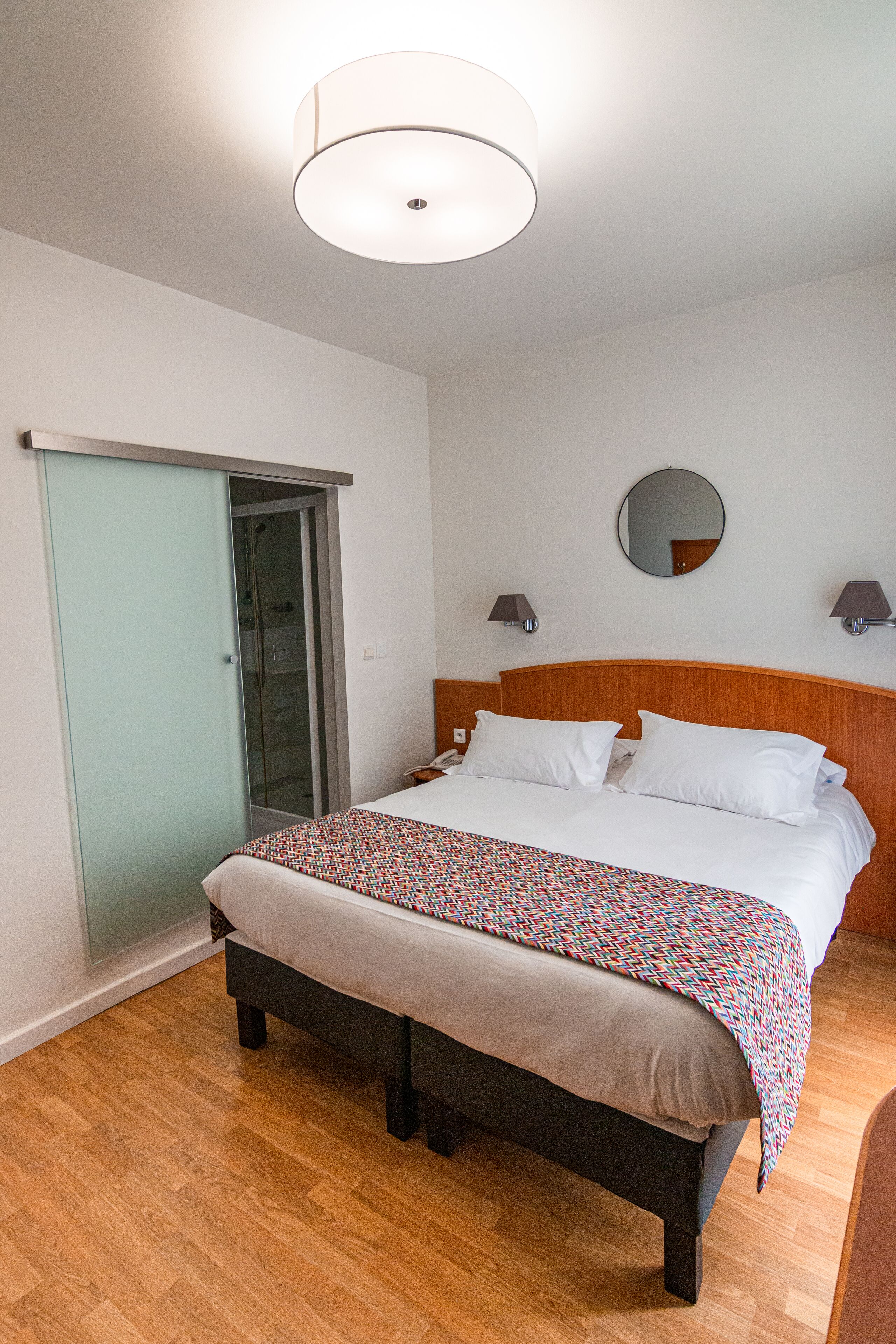 A hotel room with a bed, a bench, a ceiling light, a mirror, and a door.