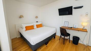 Quarto de hotel com cama, escrivaninha, cadeira, televisão e lixeira.