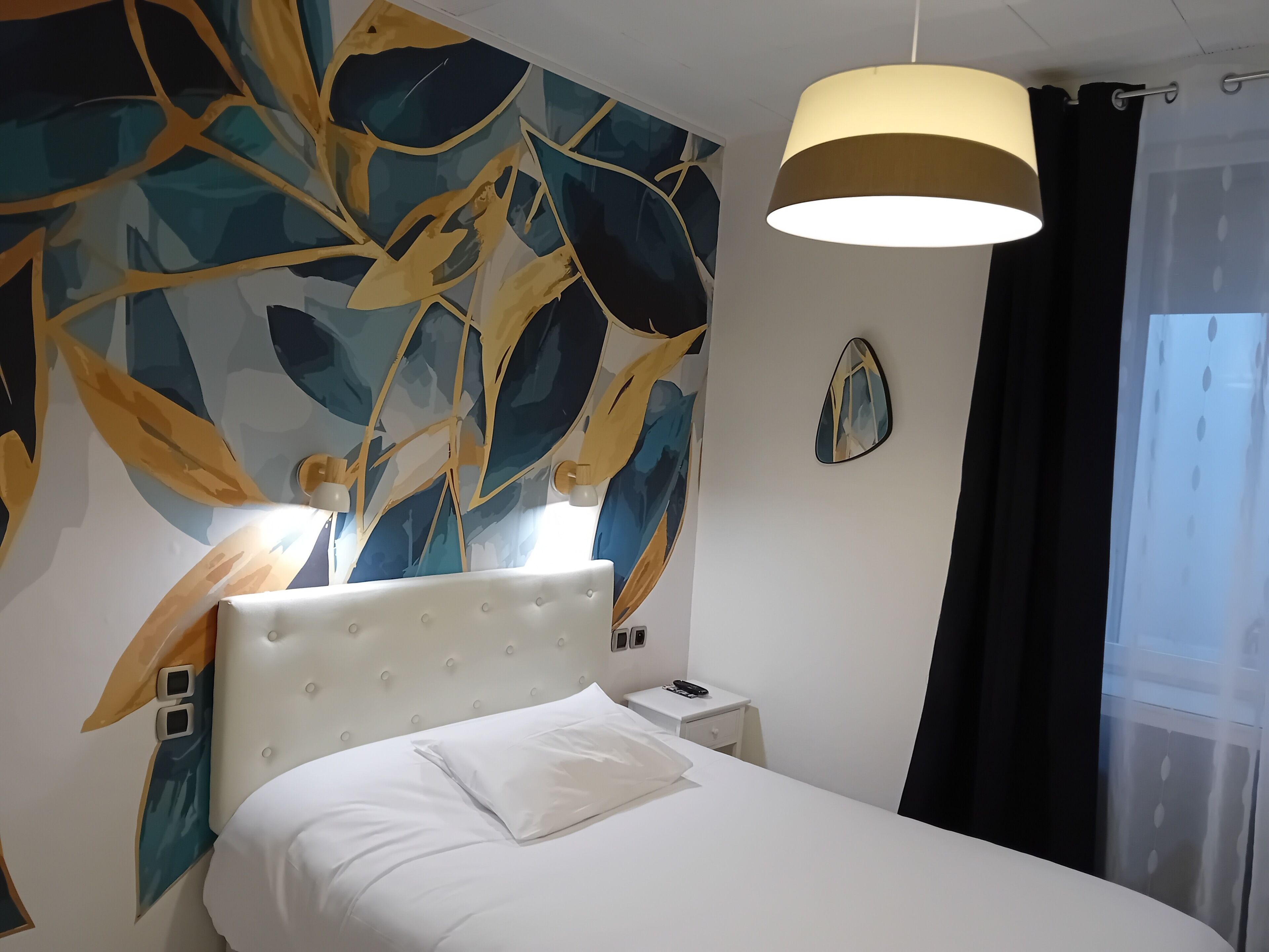 Une chambre d’hôtel avec un lit, une table de chevet, une lampe fixée au mur et une fenêtre avec des rideaux.