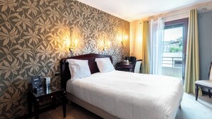 Hotelkamer met een groot bed, nachtkastje, bureau, stoel, raam met gordijnen en behang met een patroon.