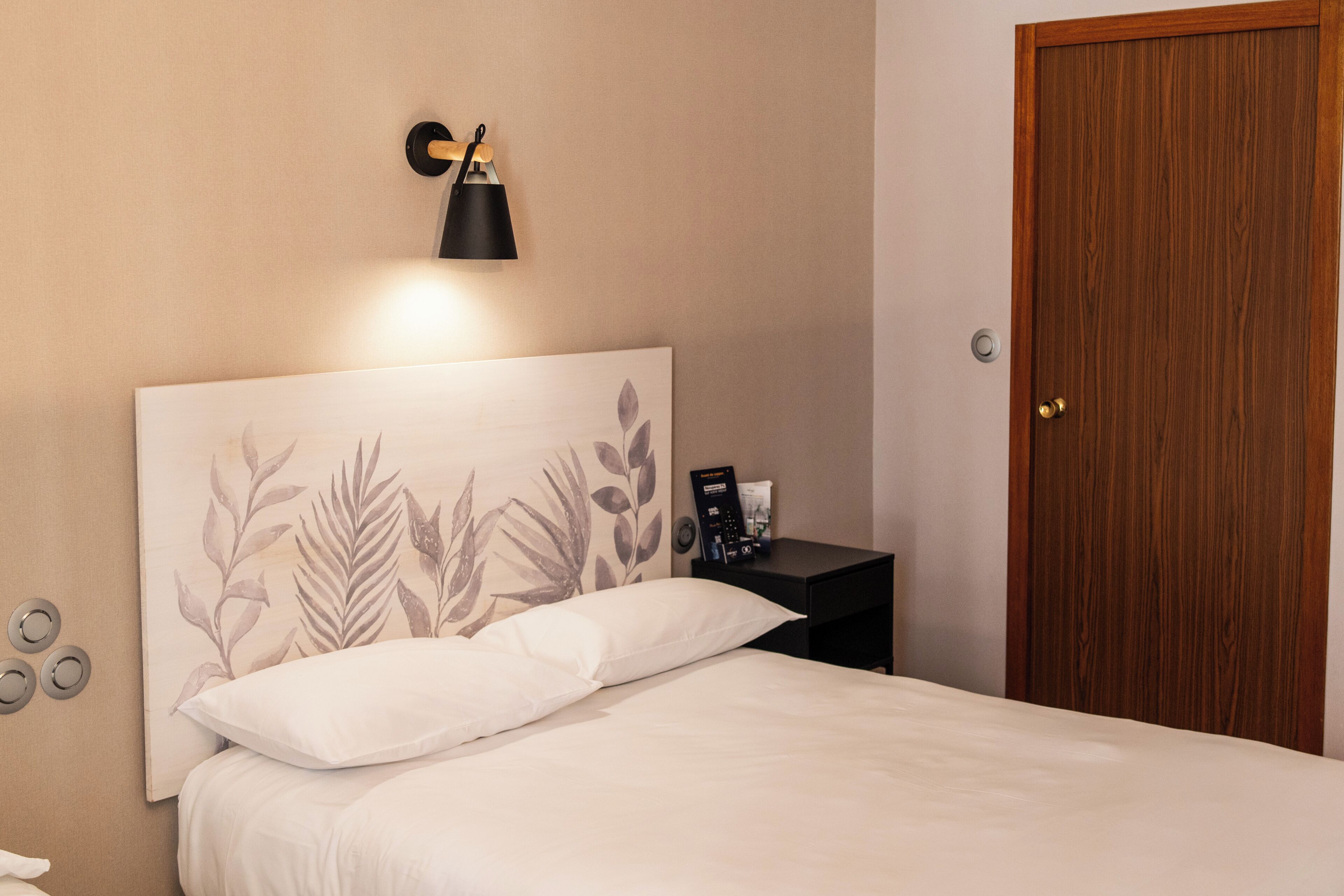 A bedroom with a bed, a bedside table with a lamp, a wall clock, and a wooden door.