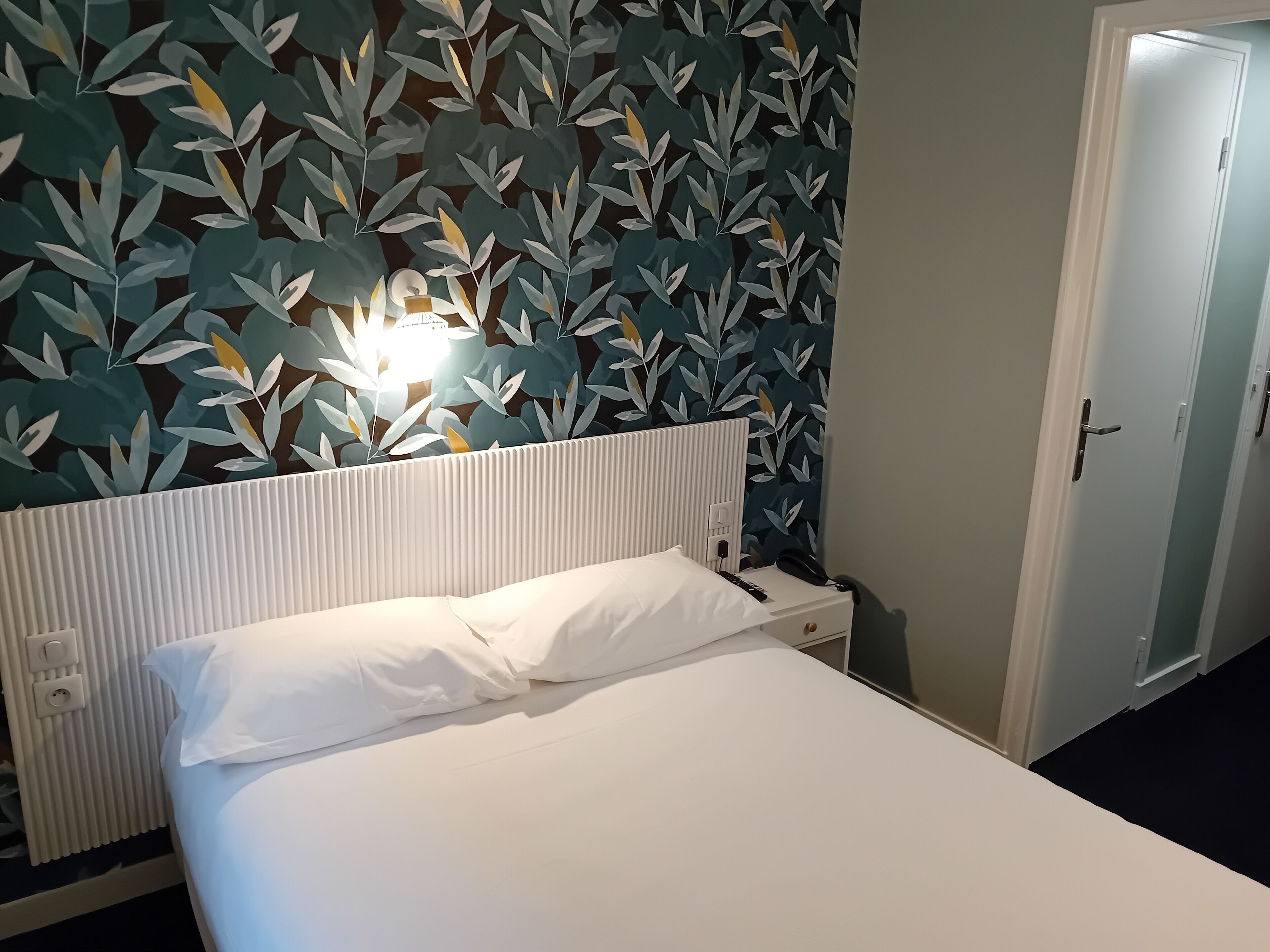 A hotel room with a bed, a nightstand, a wall-mounted lamp, and a door.