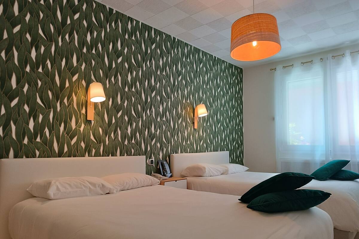 Une chambre d’hôtel avec deux lits, un mur vert à motifs, deux lampes fixées au mur et une fenêtre avec des rideaux.