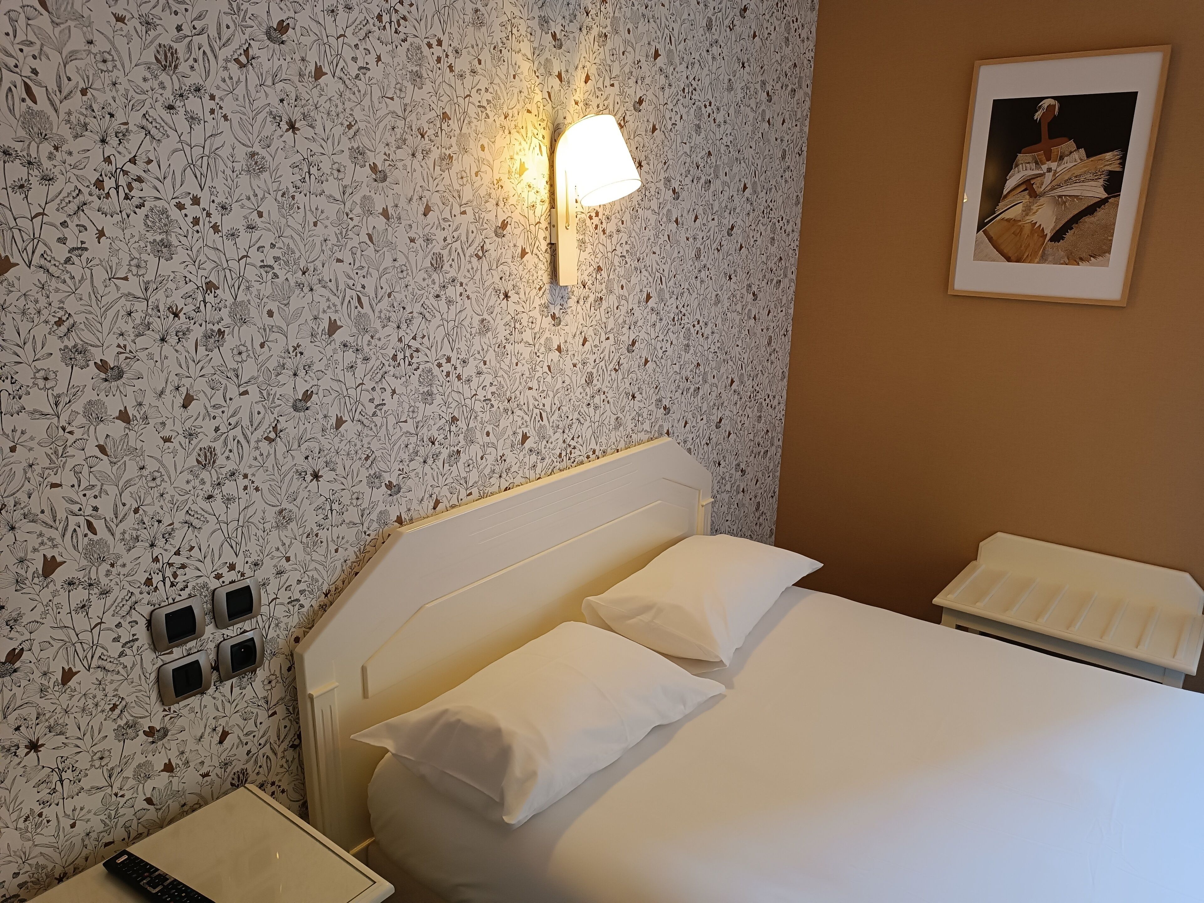 A hotel room with a floral wallpaper, a bed with white linens, a bedside table with a remote control, and a wall-mounted light fixture.