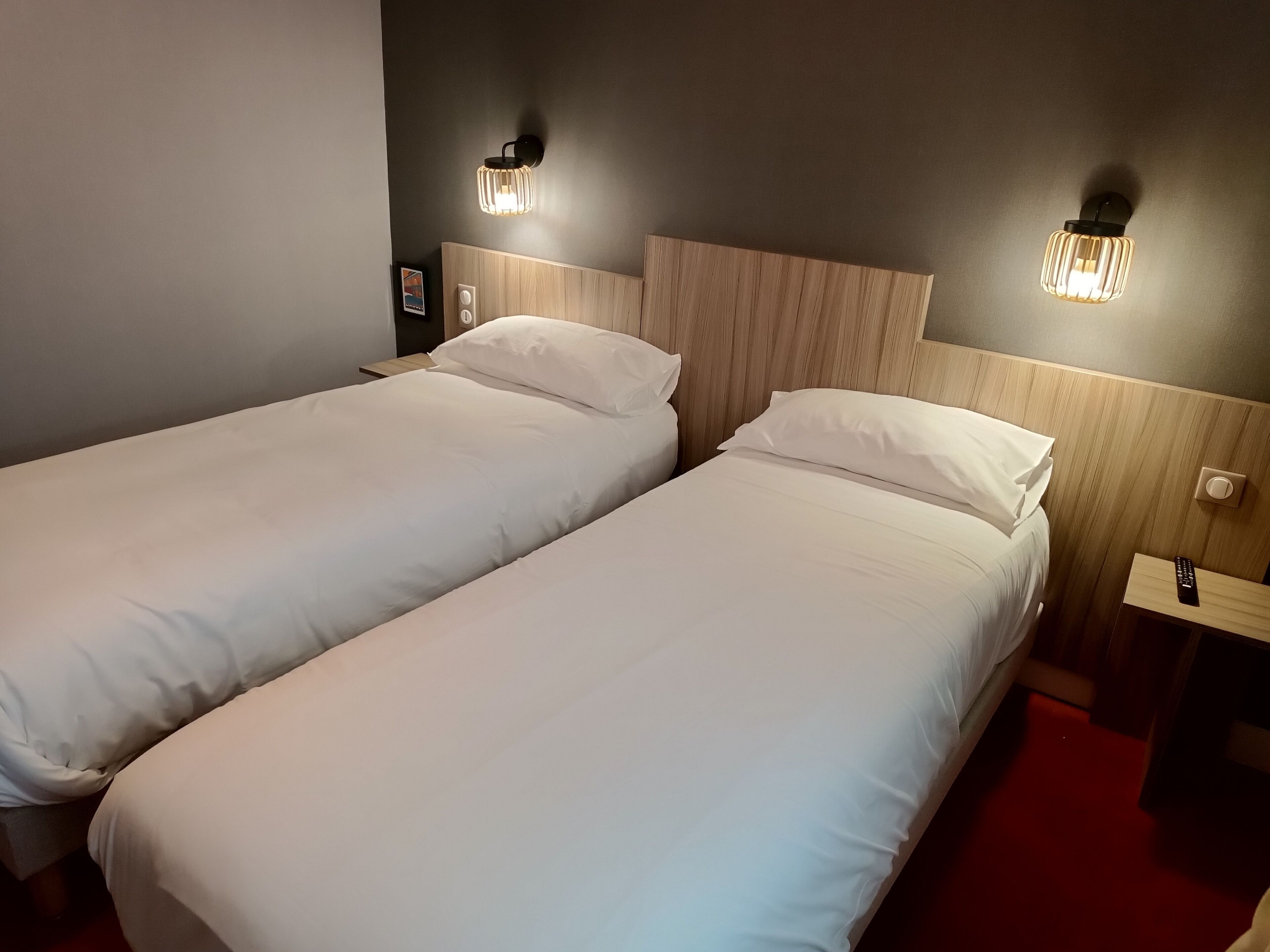 A hotel room with two single beds, a wooden headboard, bedside tables with lamps, and a red carpet.