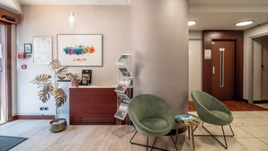 Een receptie met een bureau, twee groene stoelen, een potplant, een wandlamp en een afbeelding van een stadsgezicht in een lijst.