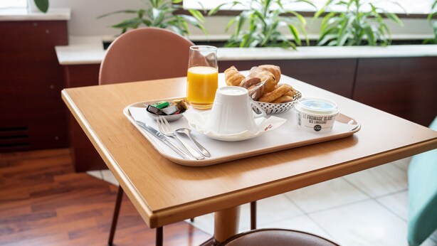 Een ontbijttray met croissants, een glas sinaasappelsap en een bakje yoghurt op een houten tafel.