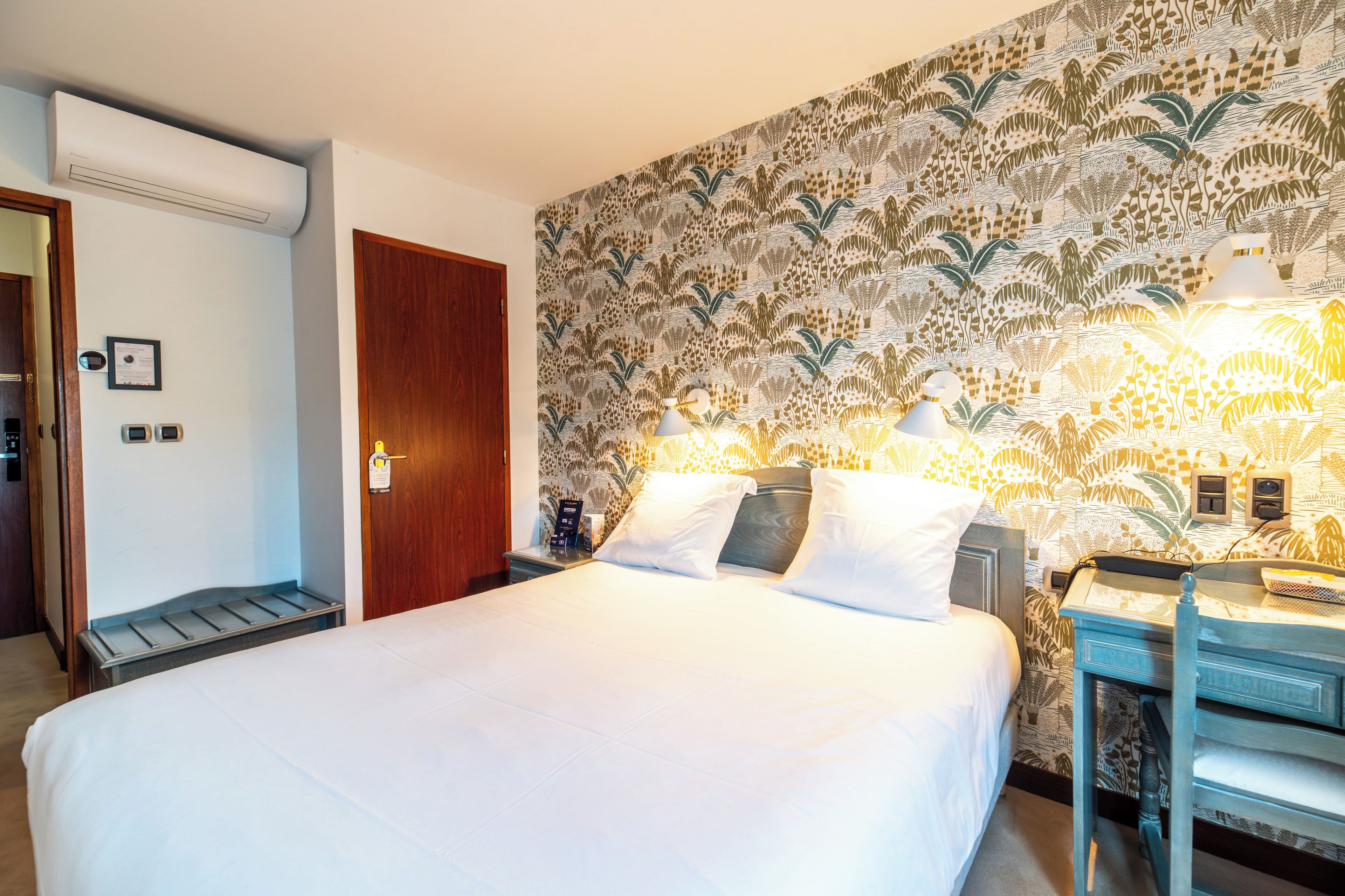 Ett hotellrum med en stor säng, ett skrivbord med en lampa, en mönstrad tapet och en dörr.