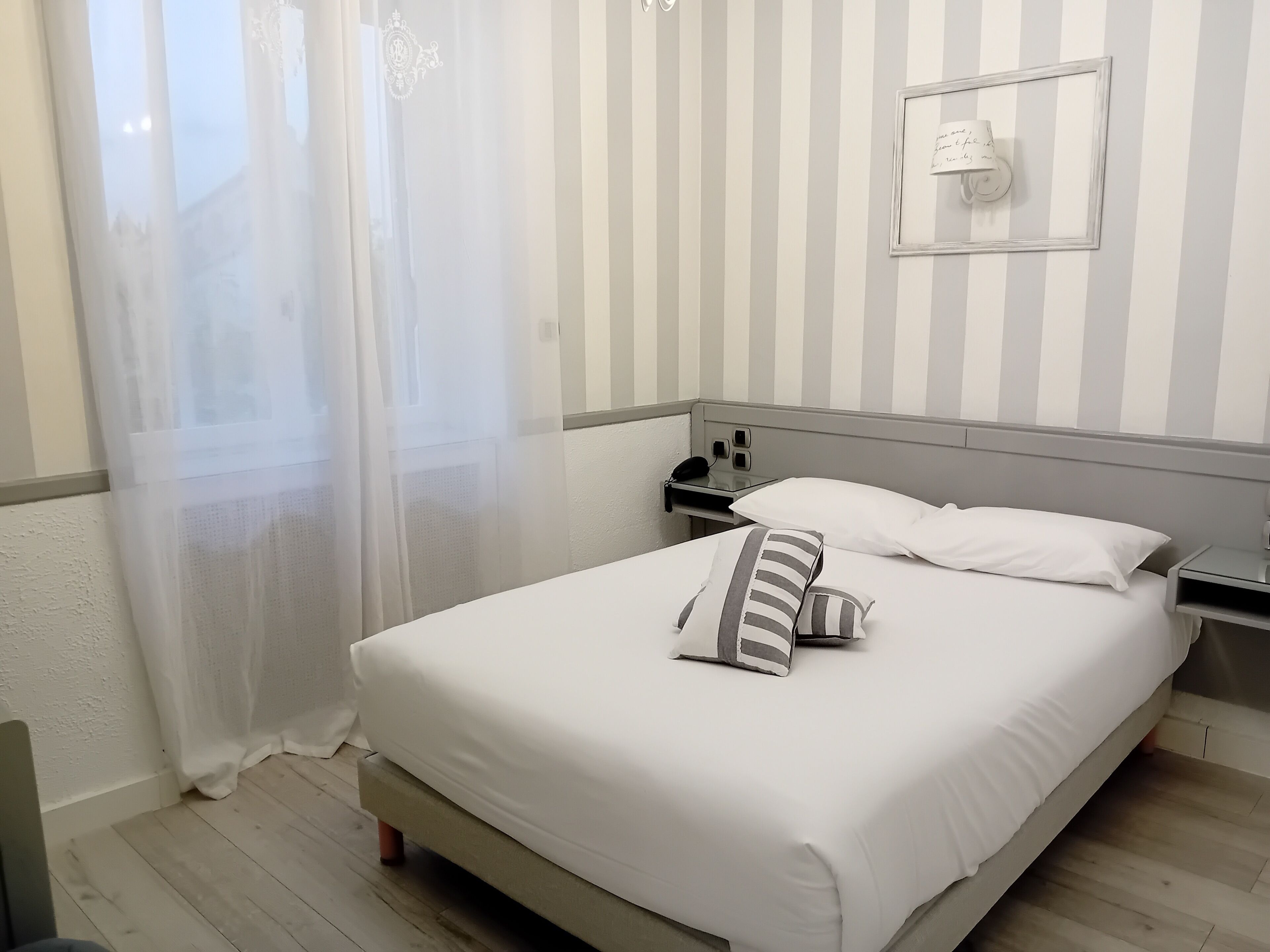 Ein ordentlich bezogenes Bett mit weißer Bettwäsche und gestreiften Kissen, ein Nachttisch mit Lampe sowie ein Fenster mit durchsichtigen Vorhängen.