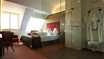 A modern hotel room with a glass-enclosed shower, a bed with white linens, a red chair, and a view of the city through a large window.