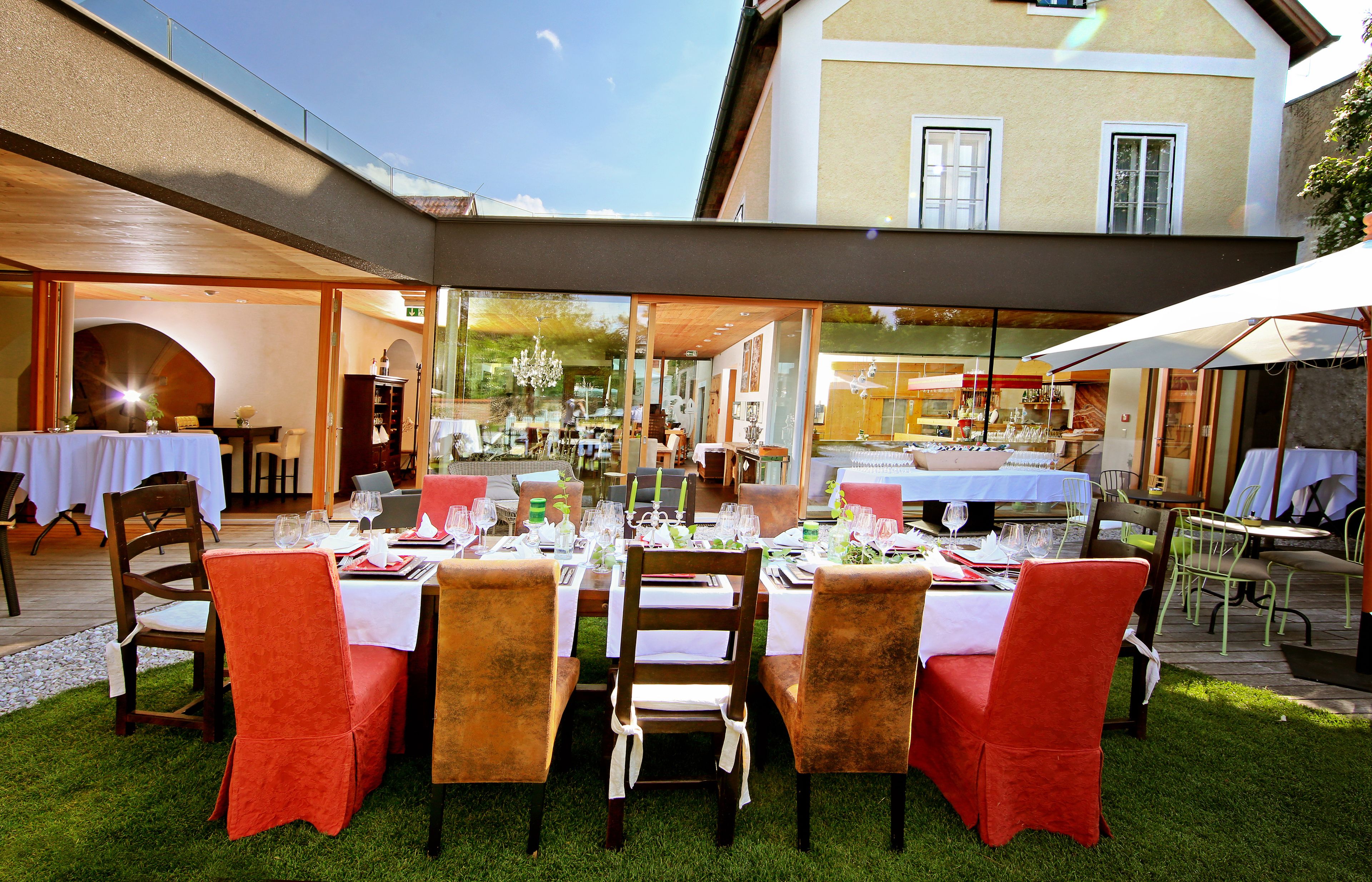 Un área de comedor al aire libre con mesas dispuestas para una comida, rodeada de sillas y un cerramiento de vidrio.