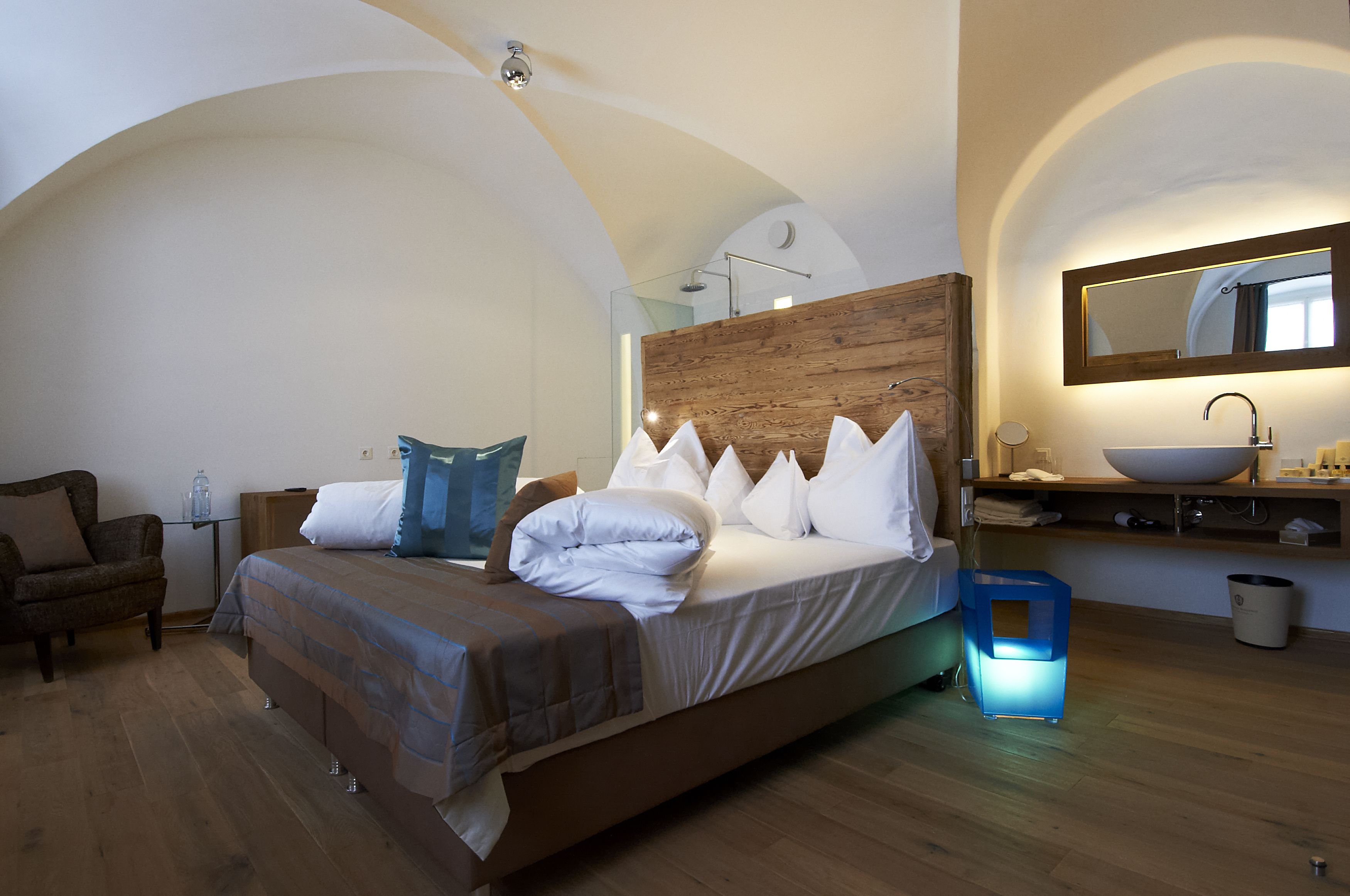A bedroom with a large bed, a wooden headboard, a bedside table, a nightstand, a mirror, a sink, and a blue bedside lamp.