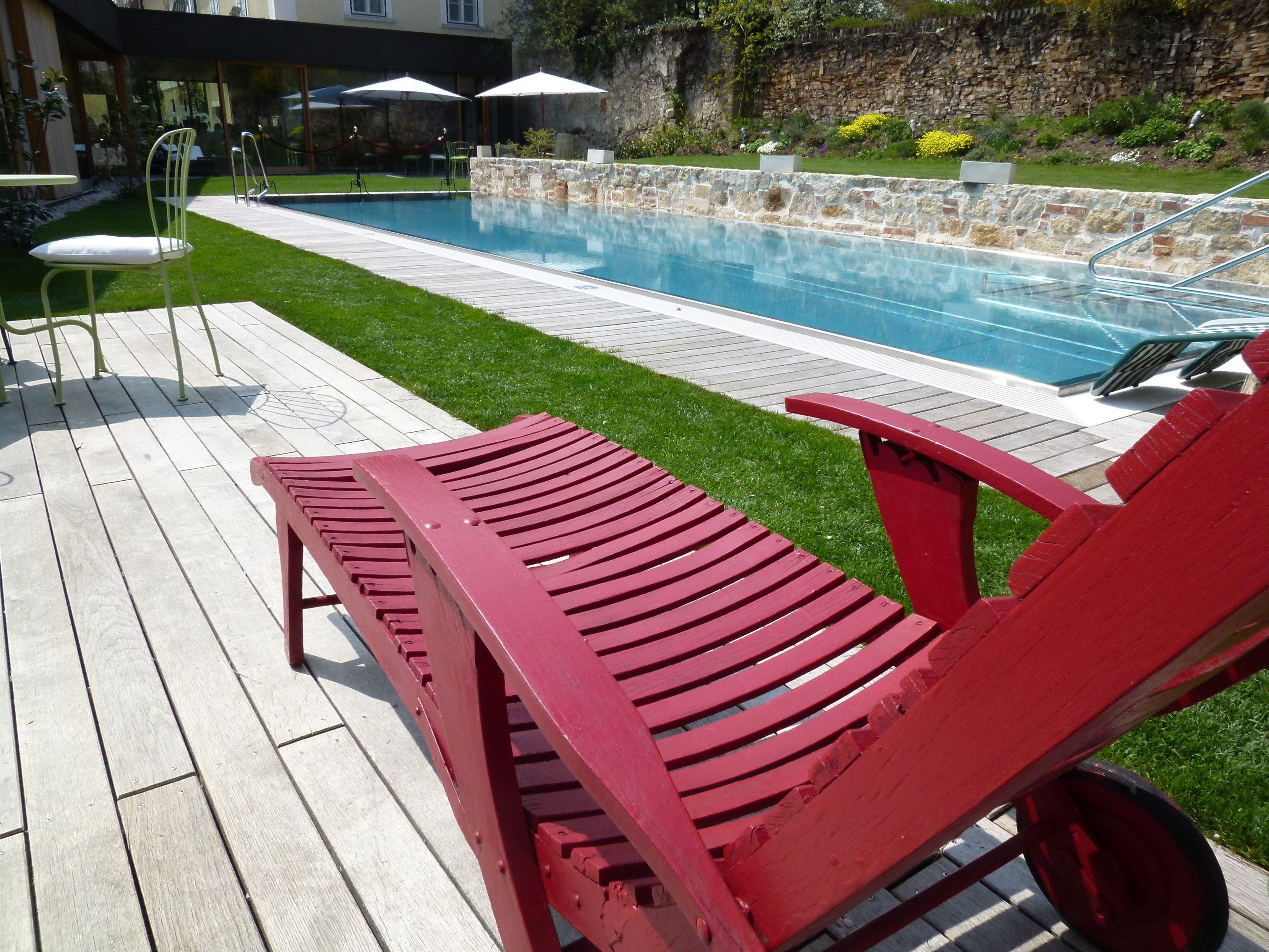 A red lounge chair by a swimming pool with a stone wall and greenery in the background.