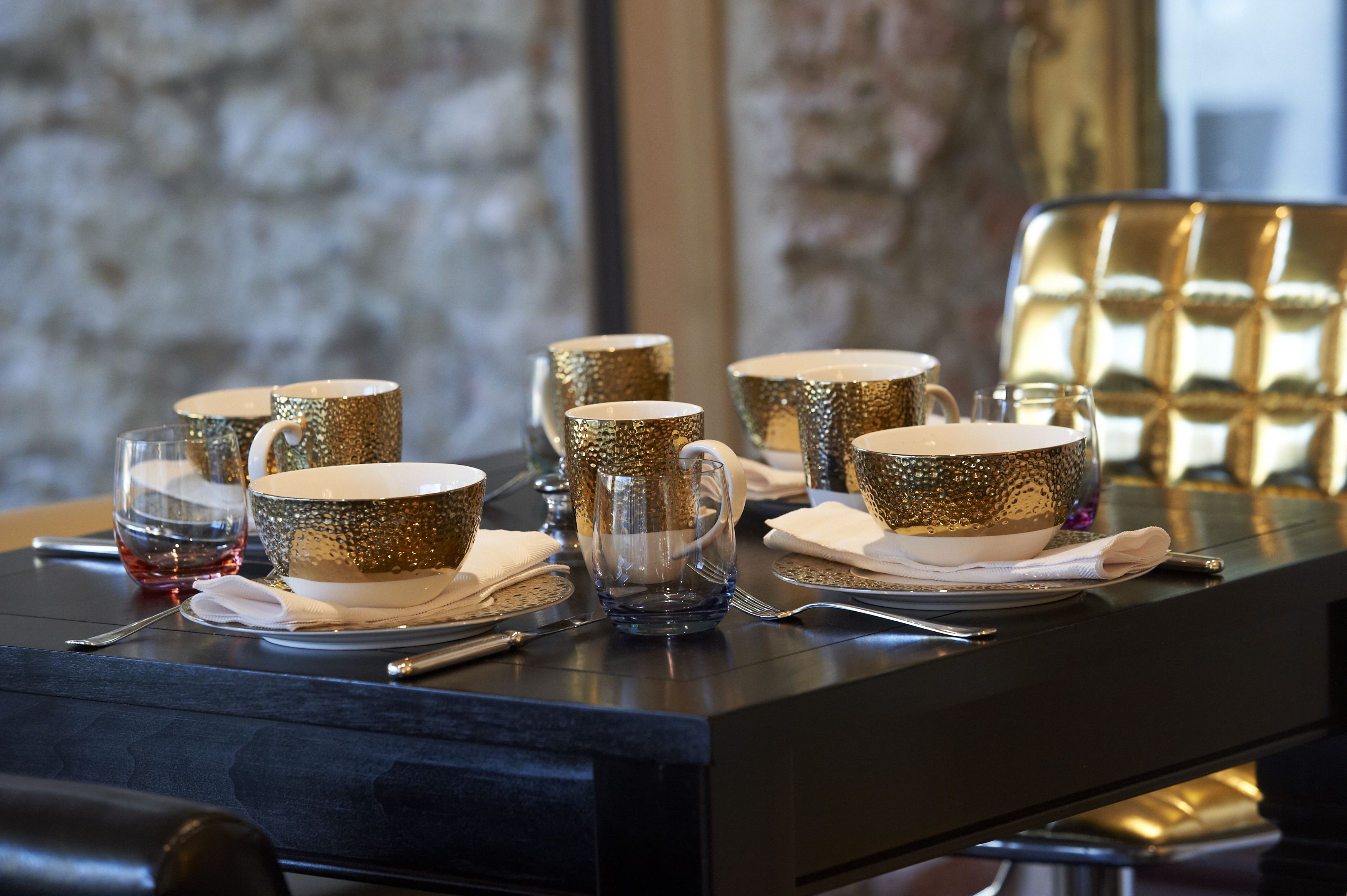 Una mesa dispuesta con cuencos dorados brillantes, vasos de vidrio transparentes y una jarra de agua, con una silla dorada al fondo.