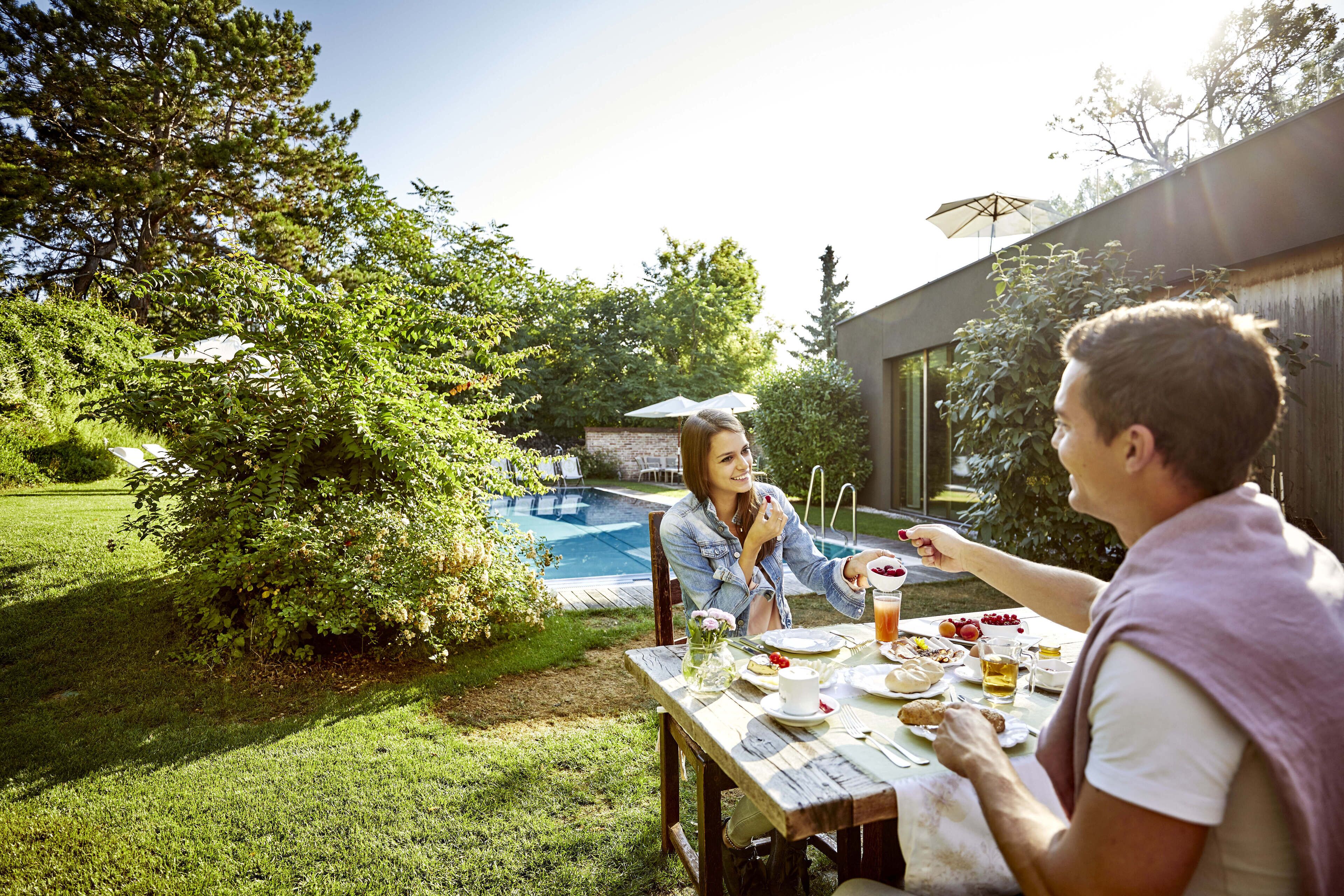 A dining table with food and drinks, surrounded by greenery and a swimming pool in the background.