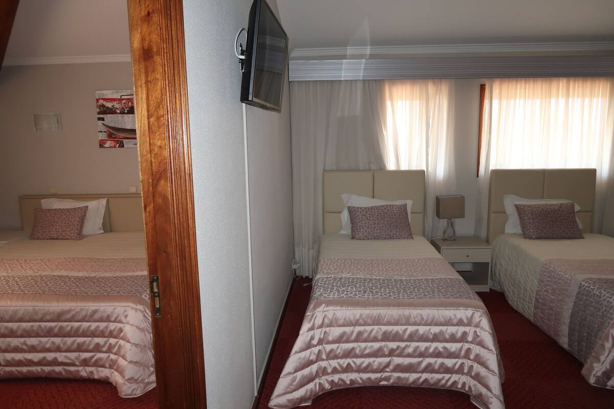 A hotel room with two beds, a TV mounted on the wall, and a window with sheer curtains.