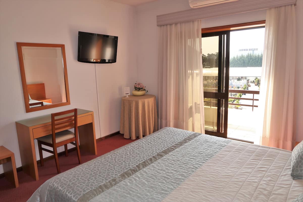 A hotel room with a bed, a desk, a chair, a television, a small table with a vase, and a balcony with a view.