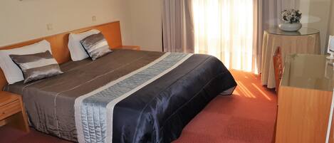 Habitación de hotel con una cama grande, una mesita de noche de madera, una pequeña mesa con un florero y un espejo.