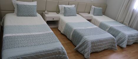 A hotel room with three single beds, each with a patterned bedspread and matching pillows. There are bedside tables with lamps and a phone.