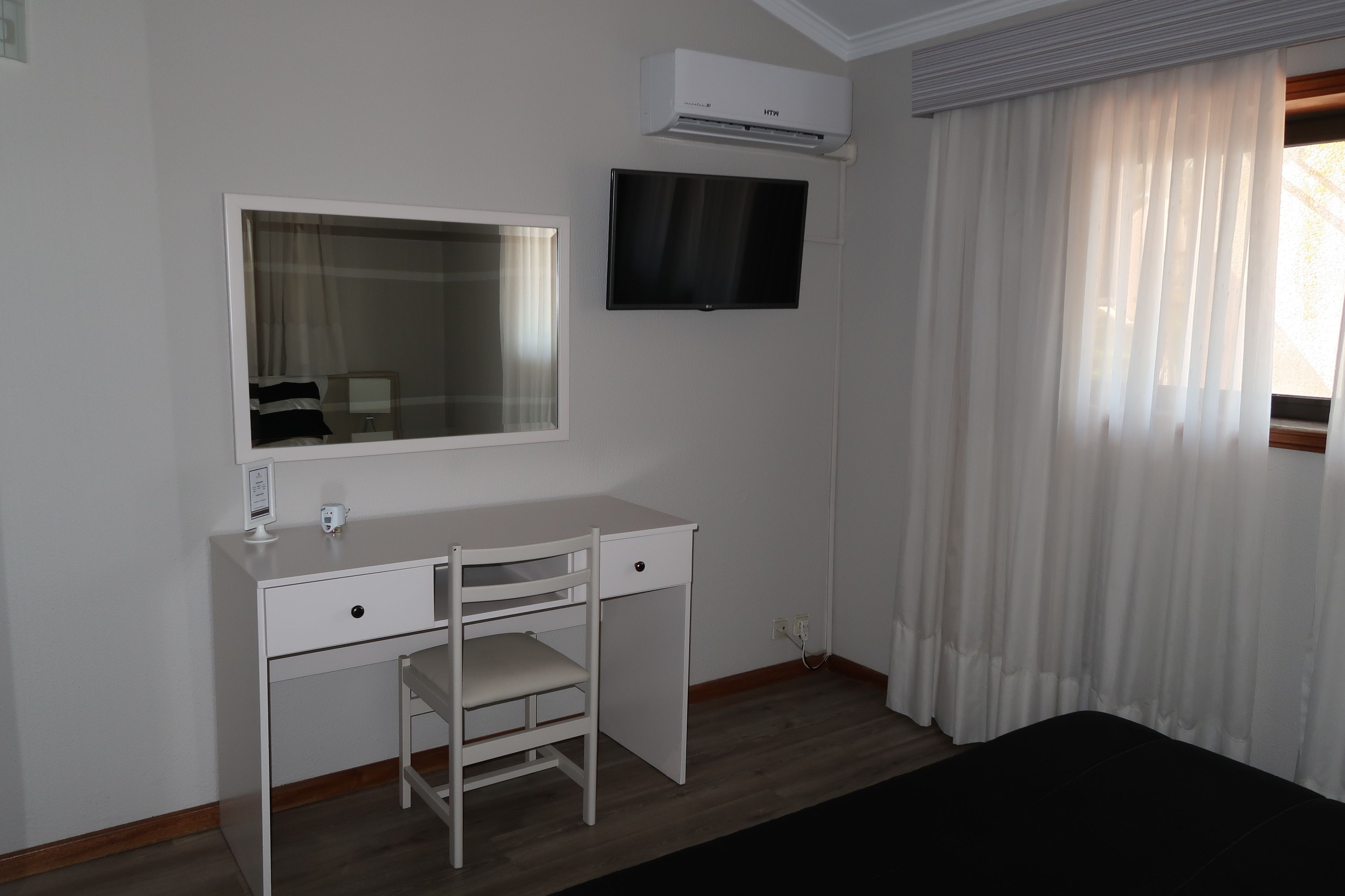Une pièce avec un bureau blanc, une chaise, un téléviseur à écran plat et une fenêtre ornée de rideaux légers.