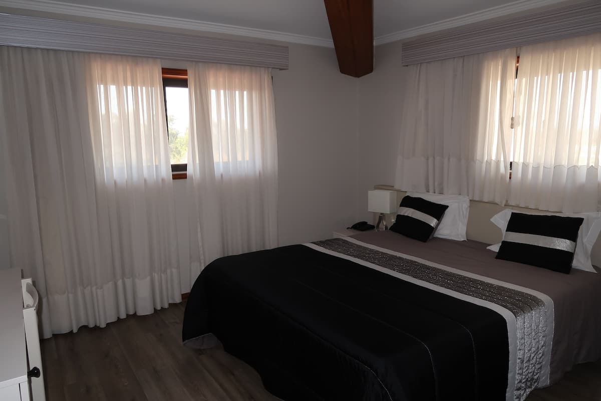 A hotel room with a large bed, white curtains, a wooden floor, and a desk with a telephone.