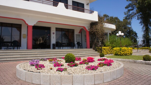 Un edificio con fachada blanca, detalles en rojo y un balcón. En la entrada hay una maceta circular con diversas flores.