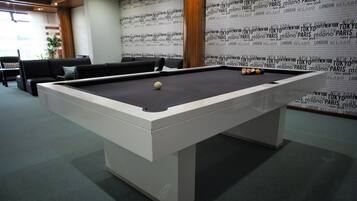Una mesa de billar en una habitación con alfombra verde, un sofá y una pared con nombres de ciudades repetidos.