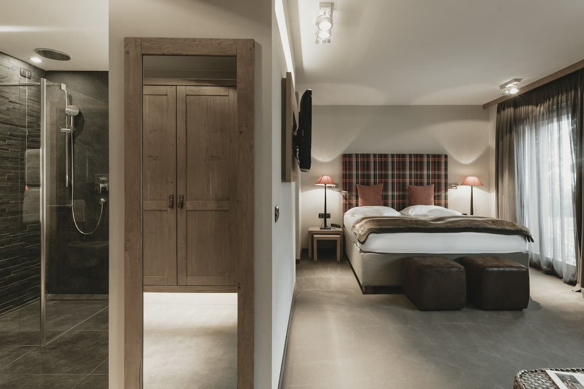 A modern bedroom with a wooden wardrobe, a glass shower enclosure, a bed with a plaid headboard, and two bedside lamps.
