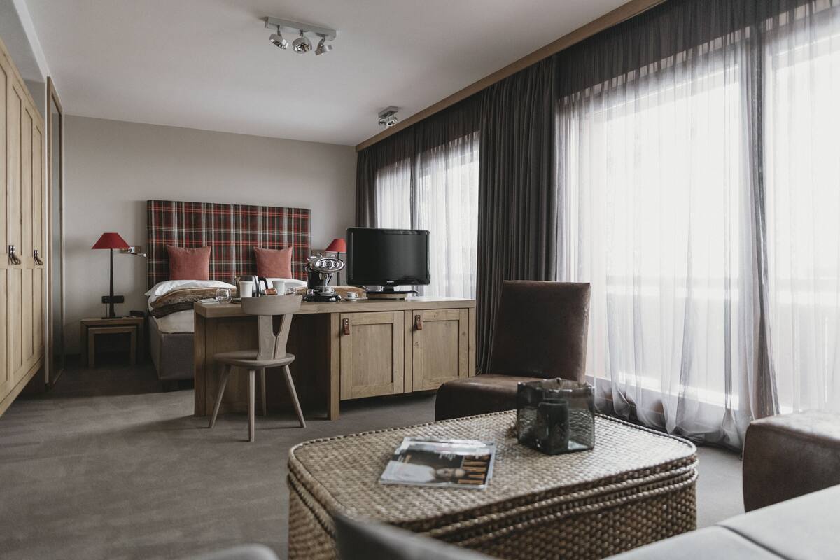 A modern hotel room with a bed, a desk with a television, a wicker coffee table, and a large window with curtains.