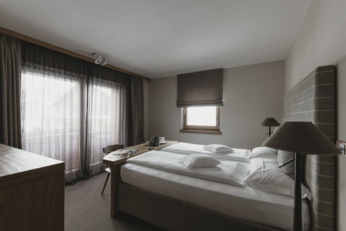 A hotel room with a bed, a desk, a chair, a lamp, a window with curtains, and a ceiling light.