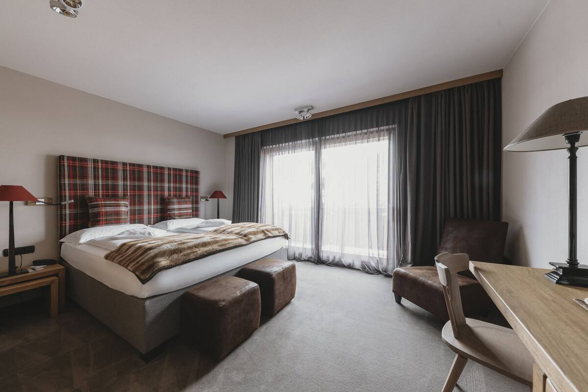 A hotel room with a bed, a desk, a chair, a lamp, and a window with curtains.