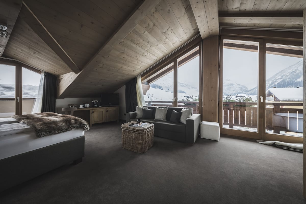 A cozy room with a bed, a sofa, and a dining area. There are large windows offering a view of snow-covered mountains.
