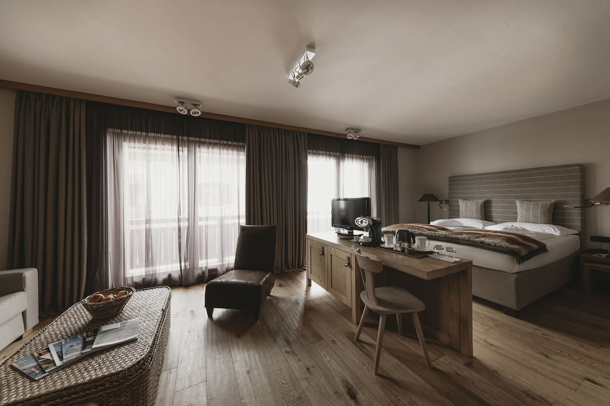 A modern hotel room with a wooden desk, a comfortable armchair, a large bed, and a view of the outdoors through sheer curtains.