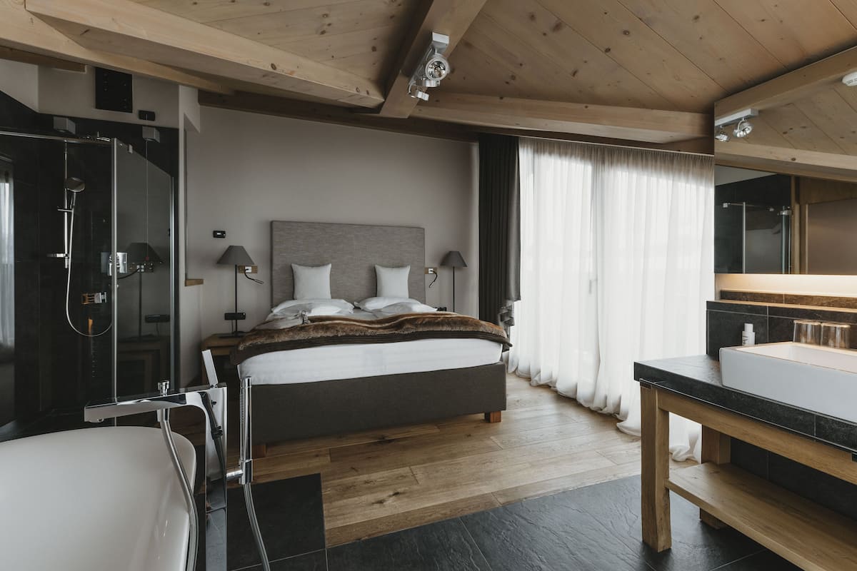 A modern hotel room with a large bed, a separate shower area, a wooden floor, and a large mirror above the sink.