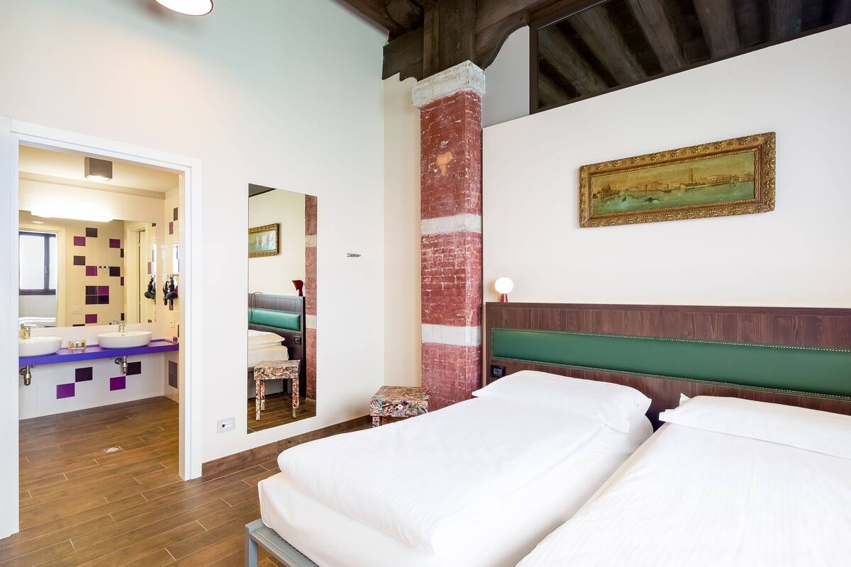 A hotel room with two beds, a large mirror, a painting, and a bathroom with a purple and white tiled wall.