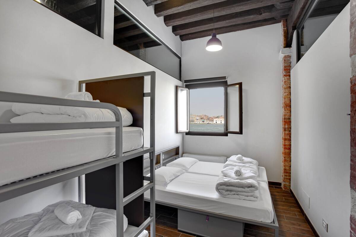 A compact hotel room with bunk beds, a window with a city view, and a brick wall.