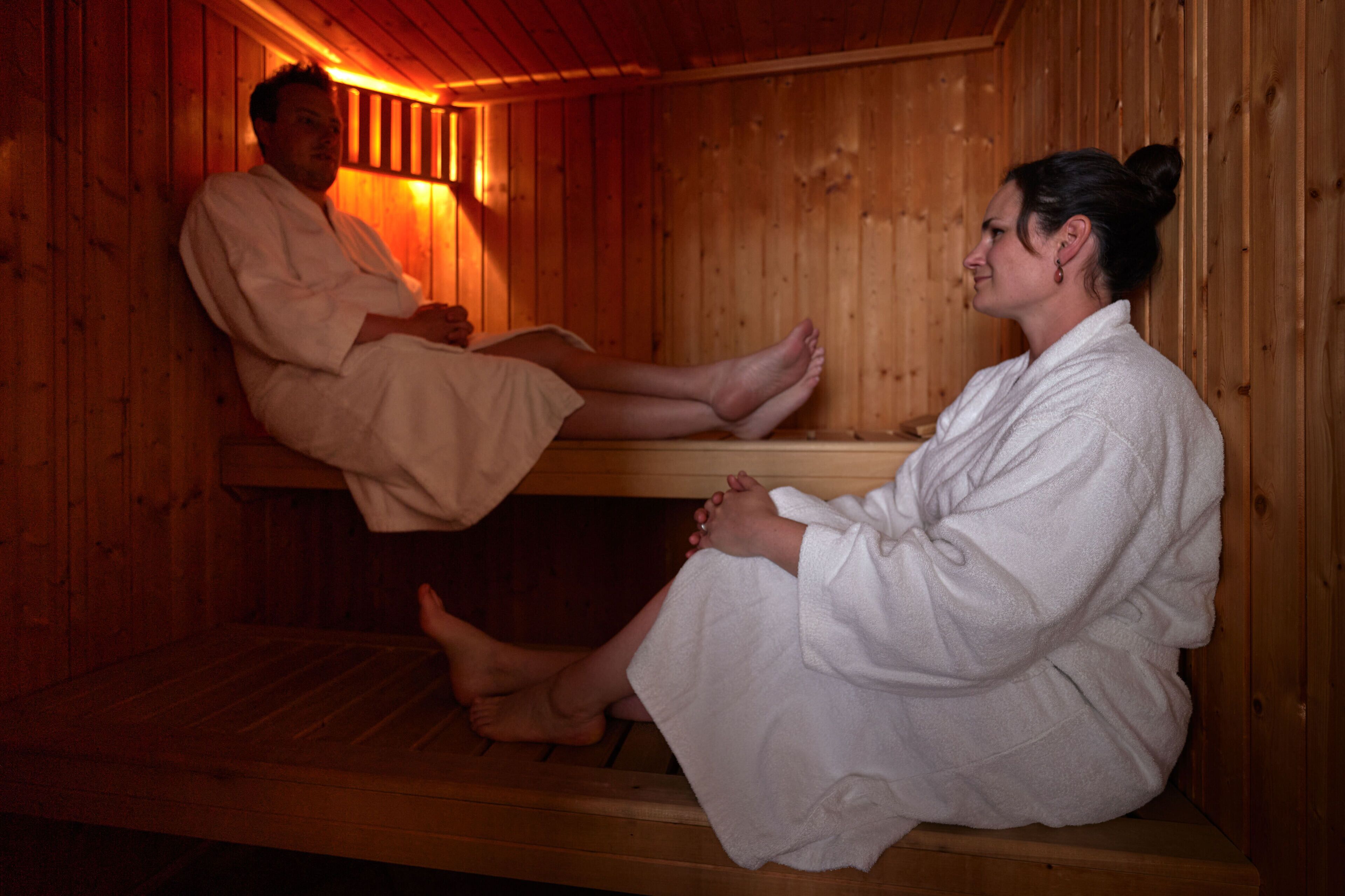 A wooden sauna with two people sitting on benches, one on the floor and one on a raised platform.