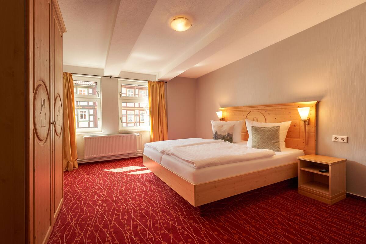 Habitación de hotel con cama de madera, mesita de noche, radiador y ventana con cortinas.