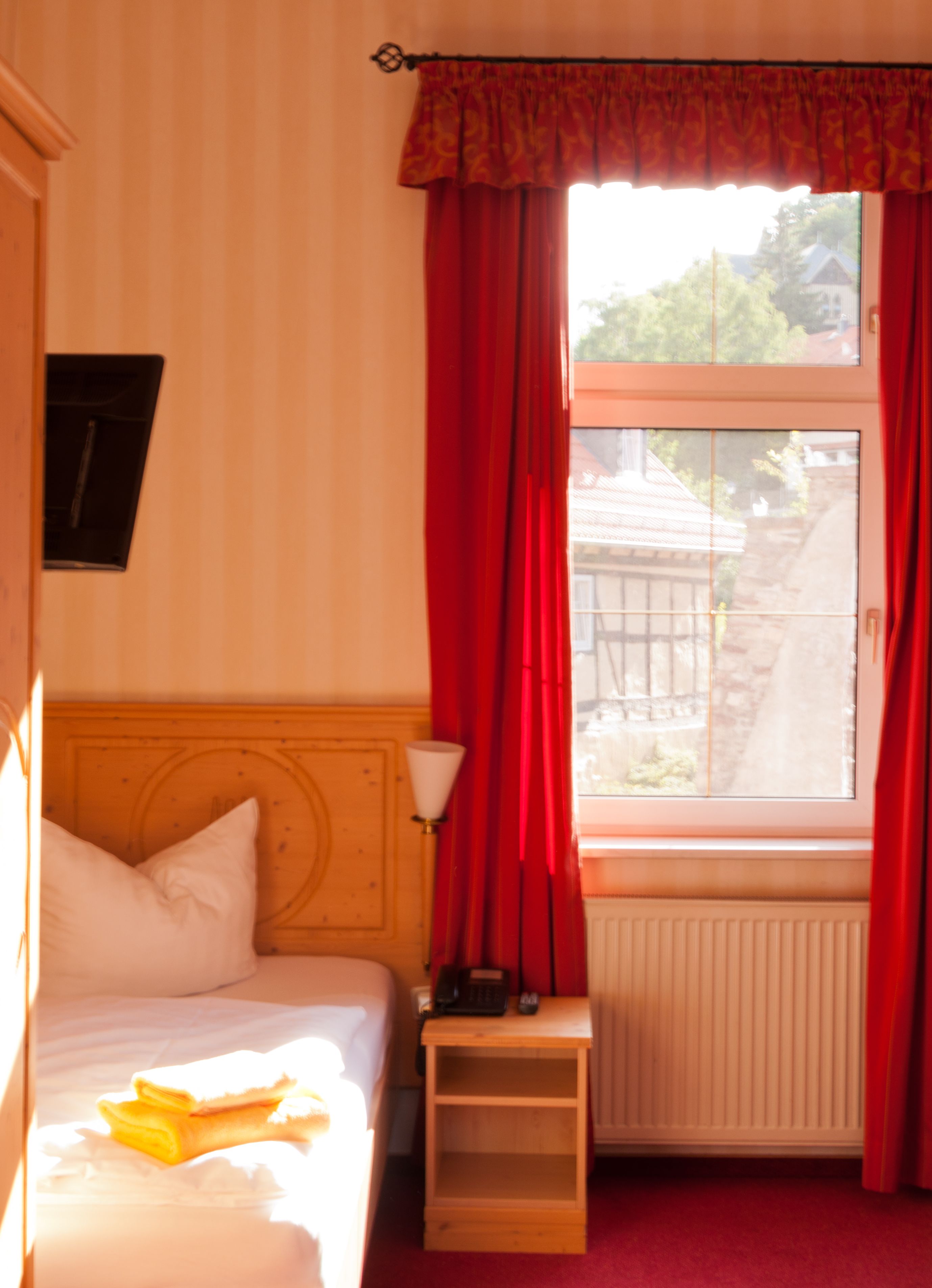 Een hotelkamer met een bed, een nachtkastje met een lamp, een raam met rode gordijnen en uitzicht op buiten.