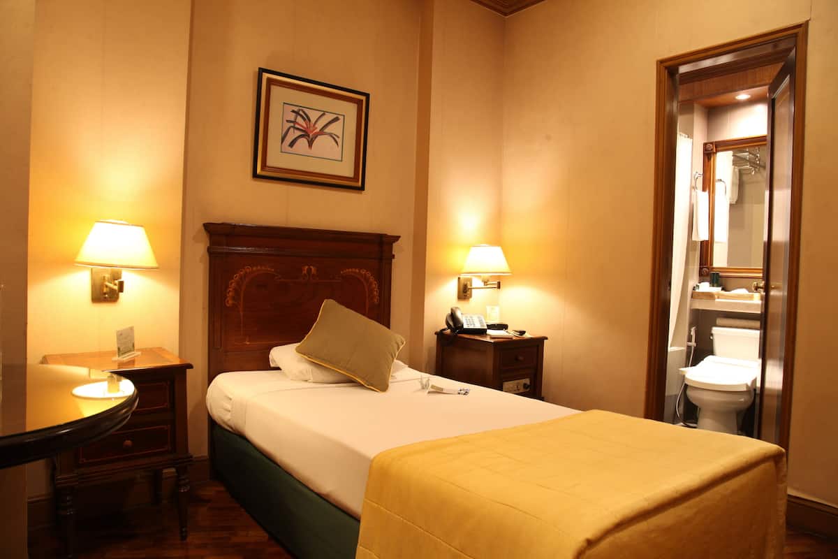 Une chambre d’hôtel avec un lit, des lampes de chevet, une tête de lit en bois, une table de chevet, un bureau et une salle de bain visible à travers une porte ouverte.